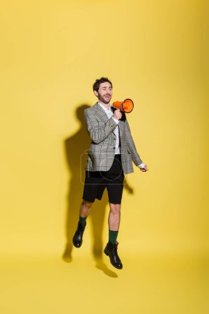 Cheerful and stylish man in shorts and jacket holding loudspeaker while jumping on yellow background 