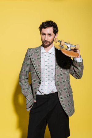 Foto de El hombre de moda en la chaqueta que sostiene el monopatín y mirando a la cámara en el fondo amarillo - Imagen libre de derechos