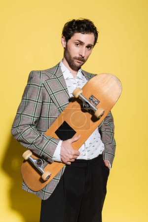 Trendy man in checkered jacket holding skateboard and posing on yellow background 