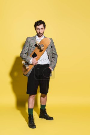 Full length of fashionable man in shorts and jacket holding skateboard on yellow background 