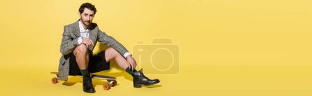 Fashionable man in jacket sitting on skateboard on yellow background, banner 