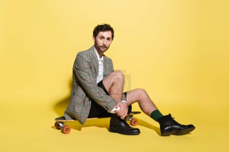 Stylish man in blazer and shorts sitting on skateboard on yellow background 