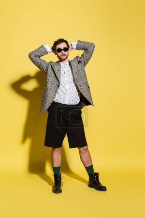 Full length of stylish model in sunglasses and blazer posing on yellow background 