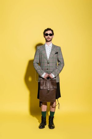 Photo for Stylish man in jacket and sunglasses holding backpack on yellow background - Royalty Free Image