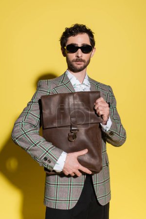Charming guy in sunglasses and blazer holding bag on yellow background 
