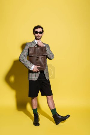 Foto de Longitud completa del modelo de moda en gafas de sol y chaqueta que sostiene el bolso marrón sobre fondo amarillo - Imagen libre de derechos