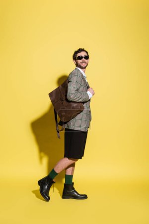 Smiling and stylish man in sunglasses posing with backpack on yellow background 