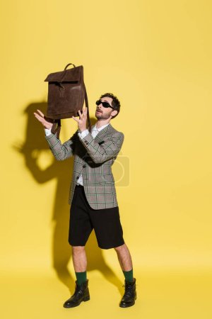 Full length of stylish man in sunglasses holding brown backpack on yellow background 