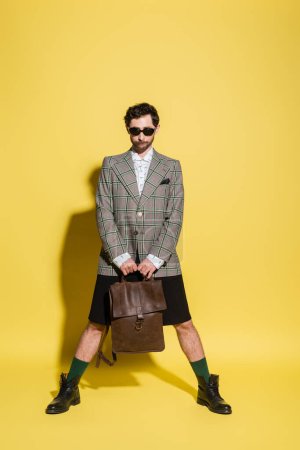 Photo for Fashionable model in jacket and sunglasses holding bag while standing on yellow background - Royalty Free Image
