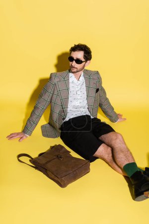 Stylish model in sunglasses sitting near brown bag on yellow background 