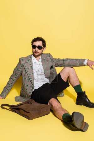 Photo for Trendy model in sunglasses and shorts sitting near bag on yellow background - Royalty Free Image