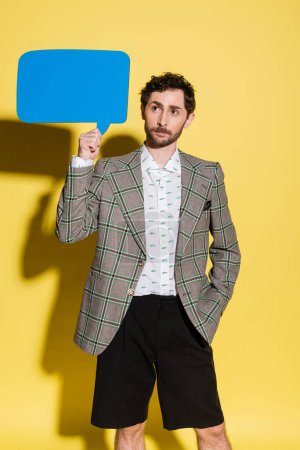 Trendy man in jacket and shorts holding speech bubble on yellow background 