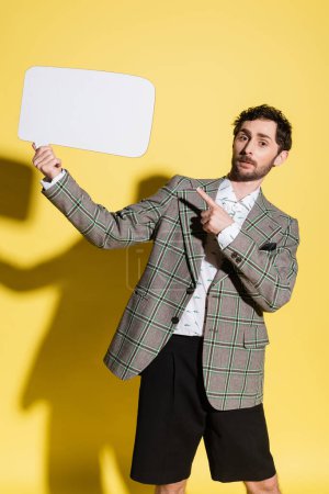 Charming guy in jacket pointing at empty speech bubble on yellow background 