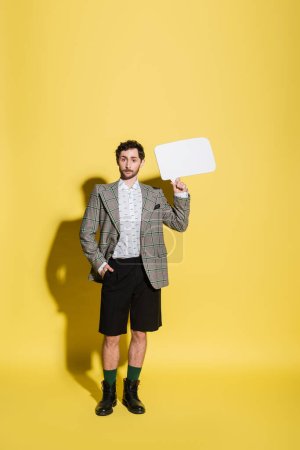 Photo for Full length of trendy model in shorts and jacket holding speech bubble on yellow background - Royalty Free Image