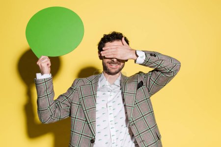 Smiling model covering eyes while holding speech bubble on yellow background 