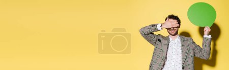 Positive model in jacket covering eyes and holding speech bubble on yellow background, banner 