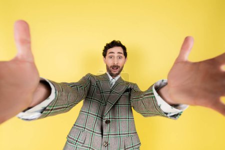 Wide angle view of stylish and excited man in jacket outstretching hands at camera isolated on yellow  