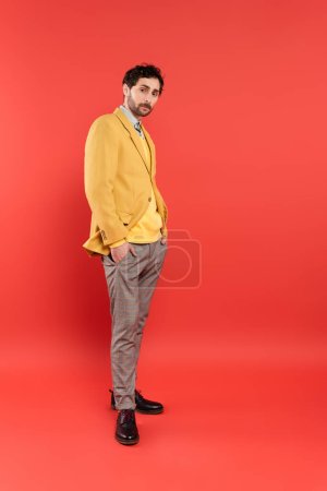 Full length of trendy model in jacket posing and looking at camera on coral red background