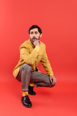 Trendy bearded man in plaid pants and yellow jacket posing on coral red background