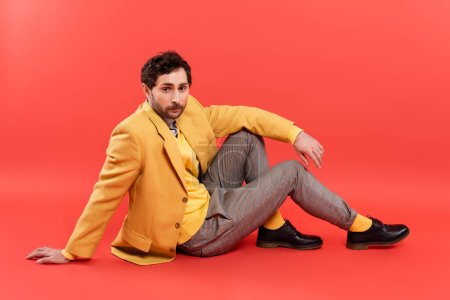 Fashionable model in yellow vest and jacket sitting on coral red background