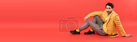 Trendy brunette man in yellow jacket sitting on coral red background, banner 