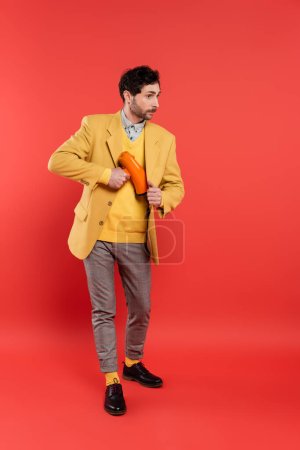 Fashionable man hiding loudspeaker under jacket on red background
