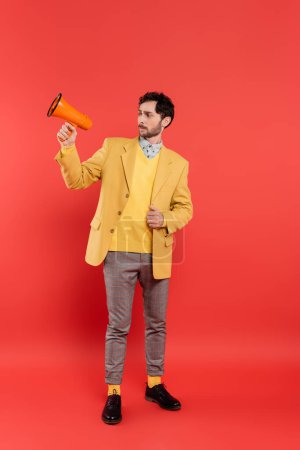 Stylish brunette man in jacket looking at loudspeaker on red background