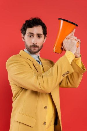 Portrait of fashionable man in jacket holding loudspeaker and looking at camera isolated on coral red 