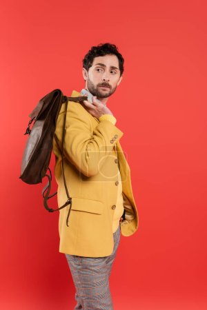 Hombre de moda con chaqueta que sostiene la mochila marrón aislada en rojo coral 