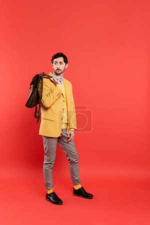 Longitud completa del modelo elegante en blazer amarillo que sostiene la mochila sobre fondo rojo coral
