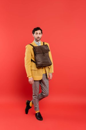 Foto de Longitud completa del modelo de moda con mochila marrón sobre chaqueta mirando a la cámara sobre fondo rojo coral - Imagen libre de derechos