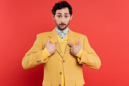 Surprised and stylish man pointing with fingers at himself on coral red background 