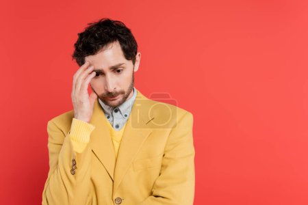 nervous man in yellow blazer touching forehead while looking down isolated on pink coral 
