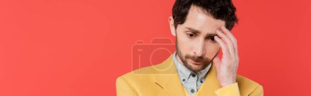 nervous man in yellow blazer touching forehead while looking down isolated on pink coral background, banner 