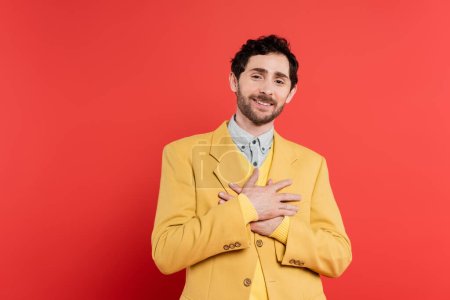 thankful man in yellow blazer holding hands near chest while standing on red coral background 