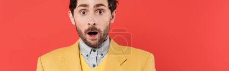 shocked man in yellow blazer standing with opened mouth and looking at camera on coral background, banner 