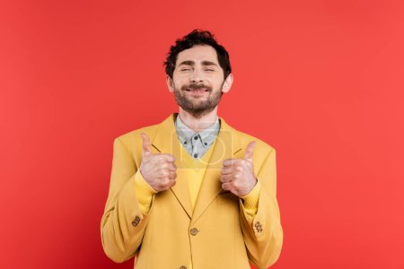 pleased man in yellow blazer standing with closed eyes and showing thumbs up on red coral background