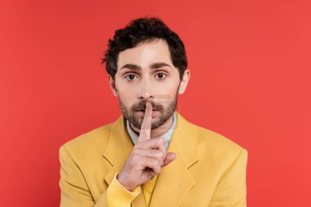 curly man in yellow blazer holding finger near lips while showing hush sign isolated on red coral 