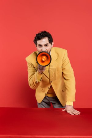 Emotionaler Mann im gelben Blazer verkündet Neuigkeiten über Lautsprecher auf rotem Korallengrund 