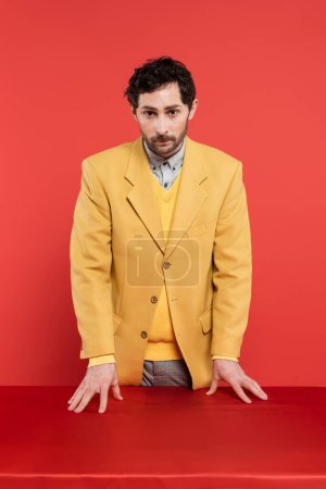 man in stylish yellow blazer standing near red desk and looking at camera on coral background 