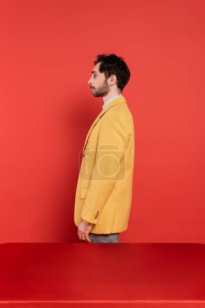Foto de Vista lateral del hombre barbudo en elegante chaqueta amarilla de pie cerca de escritorio rojo sobre fondo de coral - Imagen libre de derechos