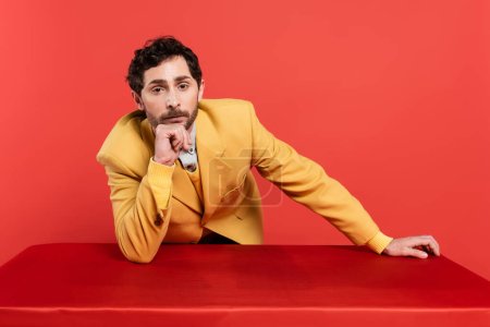 pensive man in stylish yellow blazer leaning on table and looking at camera isolated on red coral background 