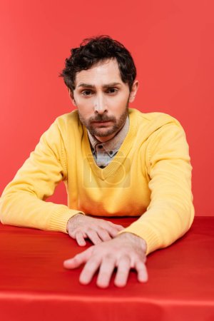 bearded guy in yellow long sleeve jumper sitting at table isolated on coral background 