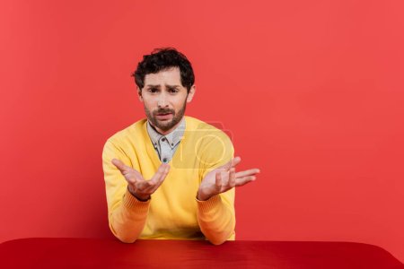 confused man in yellow long sleeve jumper sitting at table and gesturing on coral background 