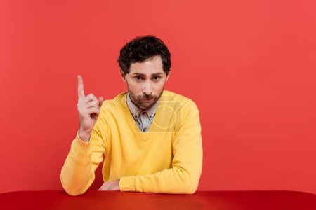 bearded man in yellow long sleeve jumper pointing with finger while sitting isolated on coral background 