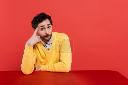 Photo for Skeptical man in yellow long sleeve jumper with finger near temple looking at camera isolated on coral - Royalty Free Image