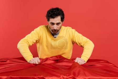stressed man in yellow jumper pulling red tablecloth on desk isolated on coral background 