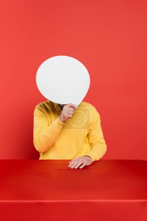 man in yellow long sleeve jumper covering face with blank speech bubble on red coral background 