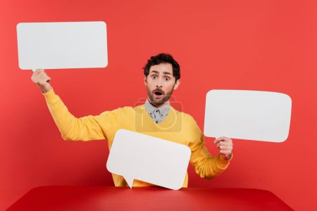shocked man in yellow long sleeve jumper holding blank speech bubbles on red coral background 