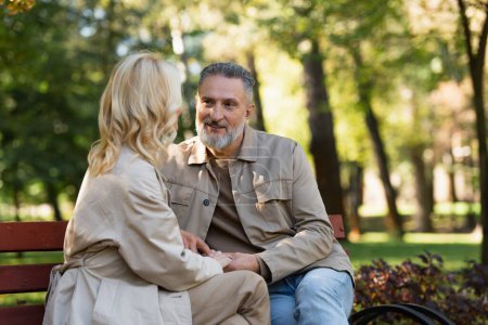 Smiling mature man holding hand of blonde wife while sitting on bench in park 